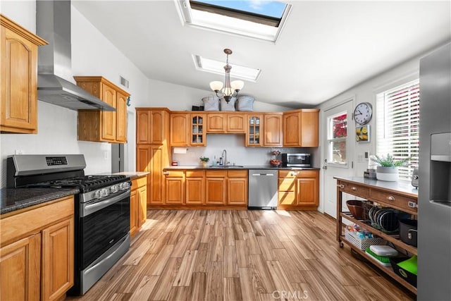 kitchen with wall chimney range hood, vaulted ceiling with skylight, glass insert cabinets, appliances with stainless steel finishes, and light wood finished floors