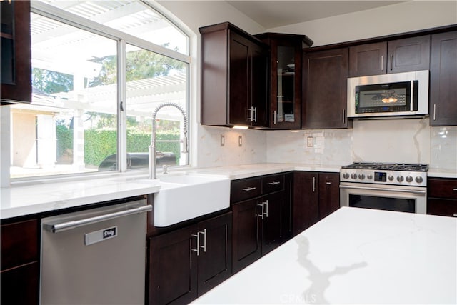 kitchen with glass insert cabinets, a sink, stainless steel appliances, backsplash, and light stone counters
