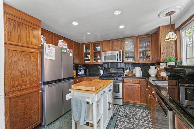kitchen with appliances with stainless steel finishes, tasteful backsplash, tile patterned floors, brown cabinetry, and glass insert cabinets