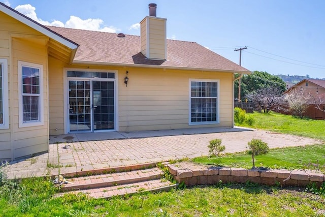 rear view of house with roof with shingles, a chimney, a patio area, and a lawn