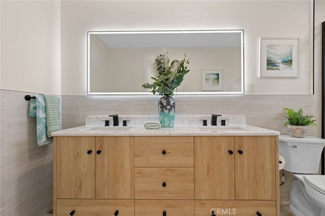 bathroom with toilet, double vanity, a sink, and tile walls
