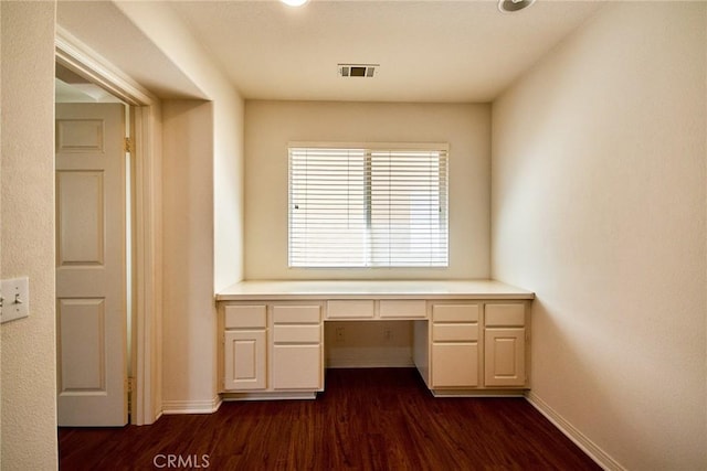 unfurnished office featuring dark wood-style flooring, built in desk, visible vents, and baseboards