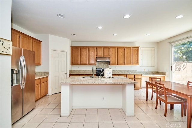 kitchen with stainless steel refrigerator with ice dispenser, an island with sink, under cabinet range hood, brown cabinetry, and recessed lighting