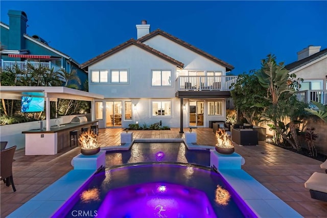 back of house at twilight featuring exterior kitchen, a chimney, a patio, stucco siding, and an in ground hot tub