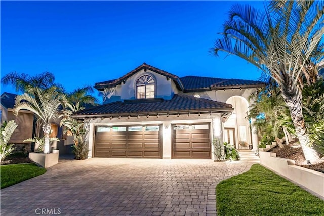 mediterranean / spanish-style home featuring a garage, stucco siding, a tiled roof, and decorative driveway