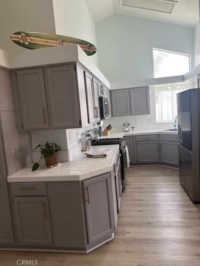 kitchen with tile counters, gray cabinets, high vaulted ceiling, light wood-type flooring, and appliances with stainless steel finishes