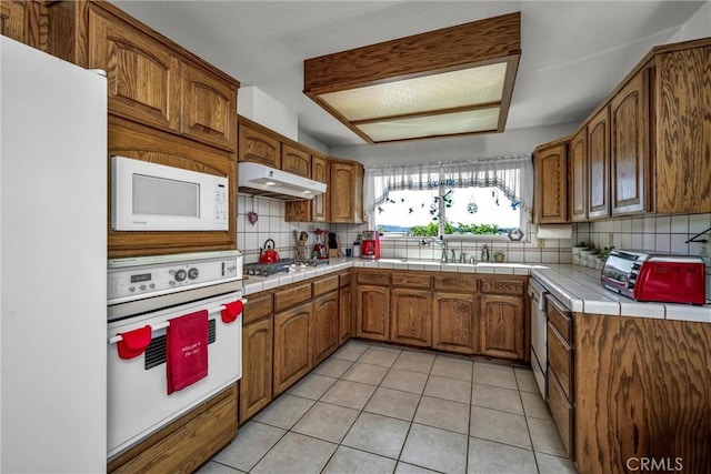 kitchen with white appliances, light tile patterned flooring, under cabinet range hood, brown cabinetry, and decorative backsplash