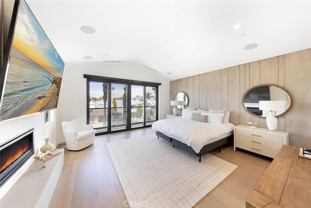 bedroom featuring wood walls, vaulted ceiling, light wood-style floors, a glass covered fireplace, and access to outside