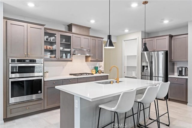 kitchen featuring a sink, under cabinet range hood, backsplash, appliances with stainless steel finishes, and an island with sink
