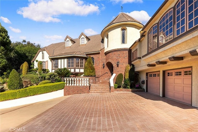 tudor home featuring a tile roof, a garage, decorative driveway, brick siding, and stucco siding