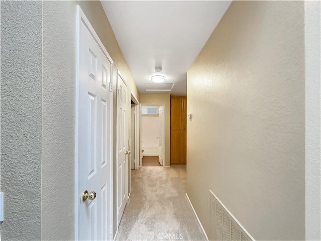 hallway with attic access, visible vents, a textured wall, baseboards, and light colored carpet