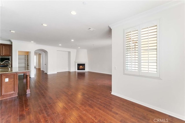unfurnished living room featuring a lit fireplace, ornamental molding, dark wood-type flooring, arched walkways, and a sink