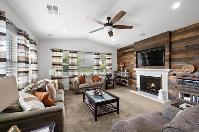 carpeted living room with a ceiling fan, vaulted ceiling, wood walls, and a lit fireplace
