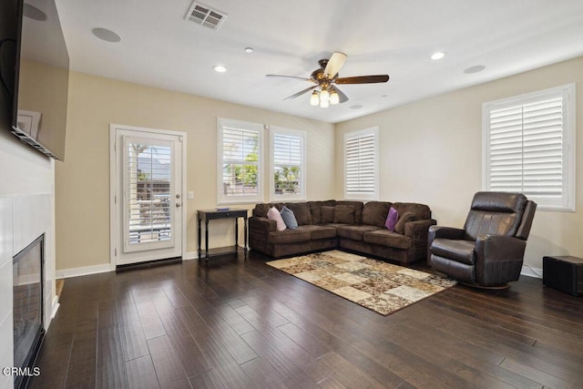 living area featuring ceiling fan, visible vents, a tiled fireplace, dark wood finished floors, and baseboards