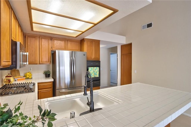 kitchen featuring tile counters, visible vents, a sink, brown cabinetry, and appliances with stainless steel finishes