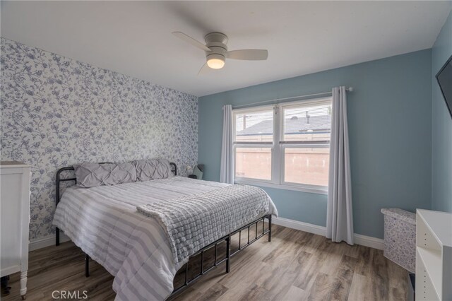 bedroom with an accent wall, wood finished floors, wallpapered walls, ceiling fan, and baseboards