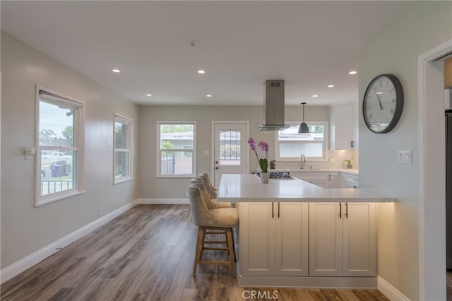 kitchen with a peninsula, a sink, island range hood, light countertops, and dark wood-type flooring