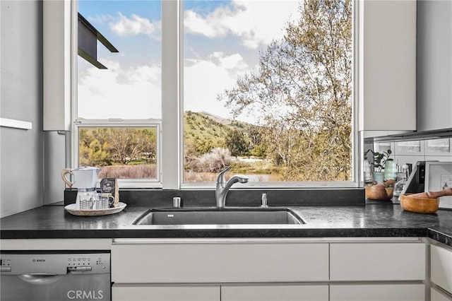 kitchen featuring dark countertops, stainless steel dishwasher, a sink, and white cabinetry
