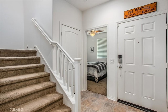 entrance foyer featuring stone finish floor, a ceiling fan, light colored carpet, baseboards, and stairs