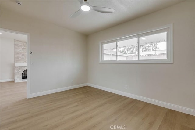 spare room with light wood-style floors, a stone fireplace, a ceiling fan, and baseboards