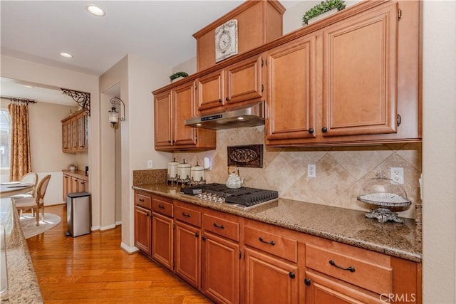 7607 Corona Valley Ave, Eastvale, CA 92880 - kitchen featuring under cabinet range hood, light wood-style floors, decorative backsplash, brown cabinetry, and stone counters