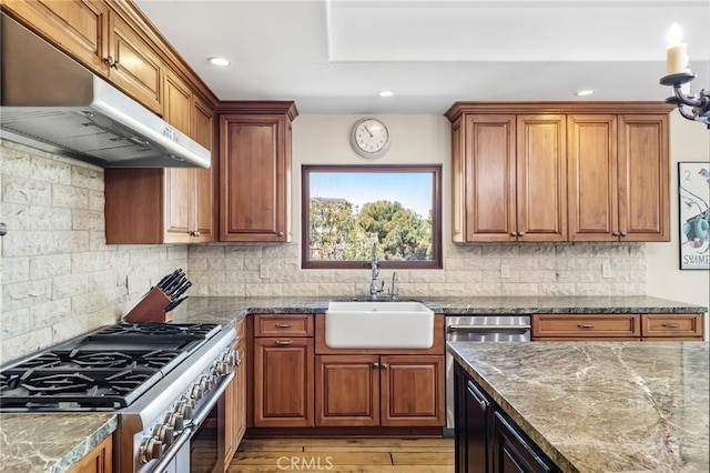 kitchen with a sink, dark stone counters, high end stainless steel range oven, brown cabinets, and under cabinet range hood