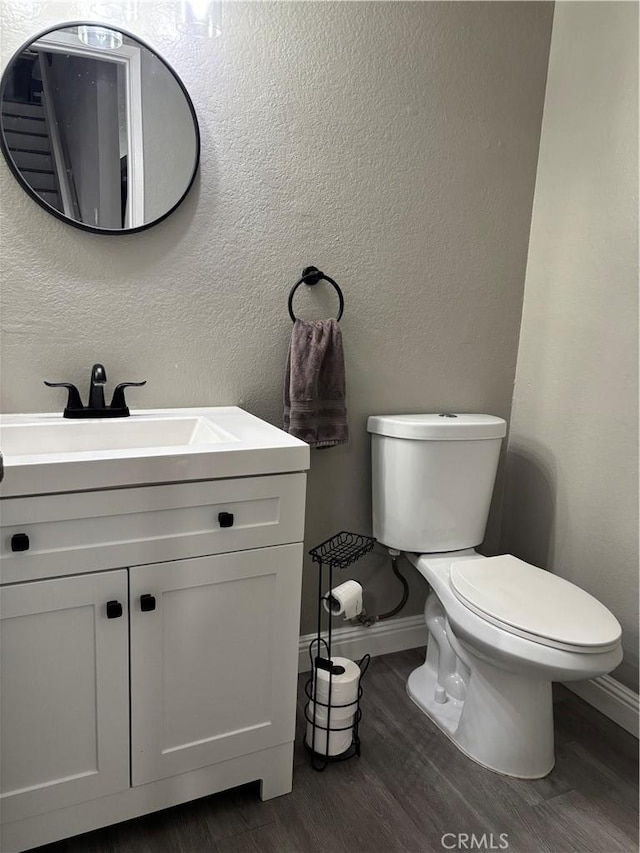 half bathroom with toilet, wood finished floors, vanity, a textured wall, and baseboards
