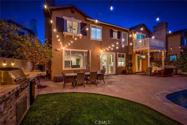 back of house at night featuring a balcony, a patio, stucco siding, an outdoor kitchen, and outdoor dining space