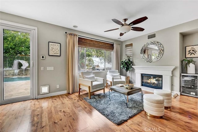 living area featuring beverage cooler, a glass covered fireplace, a ceiling fan, wood finished floors, and visible vents