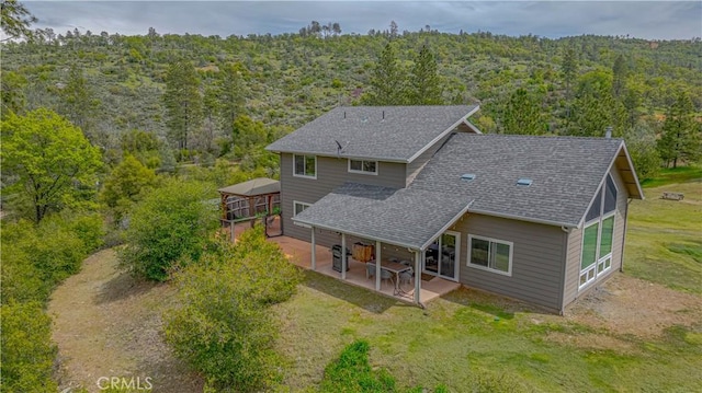 back of property with a lawn, roof with shingles, a patio, and a view of trees