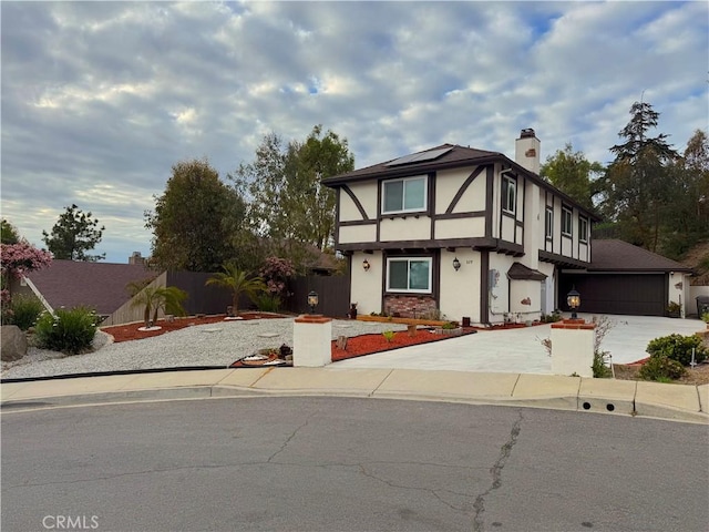 tudor-style house with a chimney, driveway, roof mounted solar panels, fence, and stucco siding