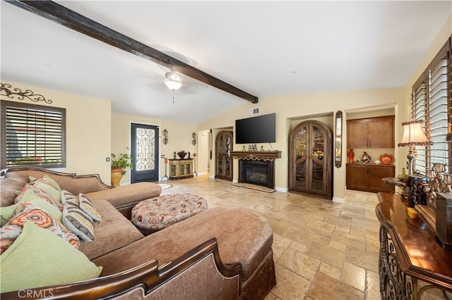 living room with a ceiling fan, stone tile floors, a glass covered fireplace, vaulted ceiling with beams, and baseboards