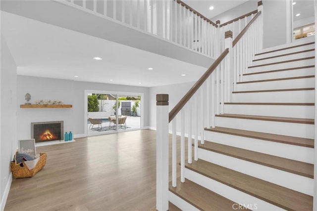 stairs featuring wood finished floors, recessed lighting, a lit fireplace, baseboards, and a high ceiling