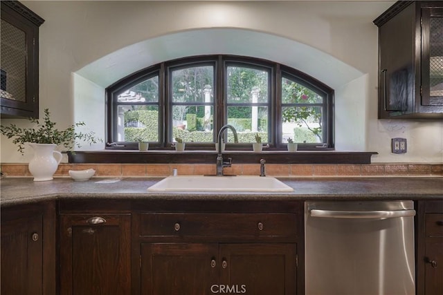 kitchen with stainless steel dishwasher, a sink, a wealth of natural light, dark brown cabinets, and dark countertops