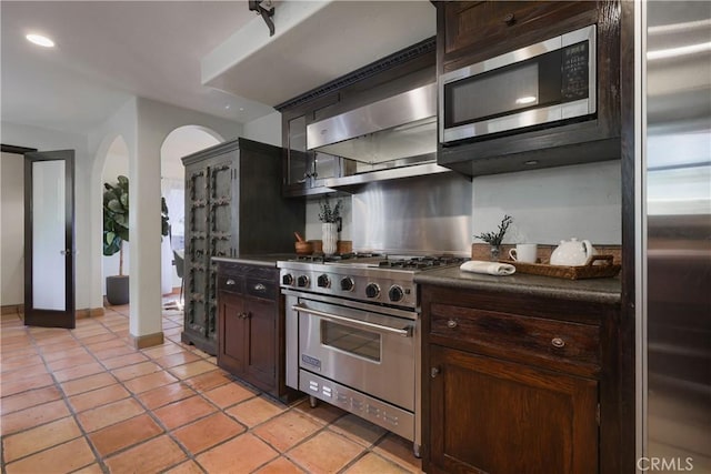 kitchen featuring stainless steel appliances, dark countertops, dark brown cabinets, arched walkways, and light tile patterned flooring