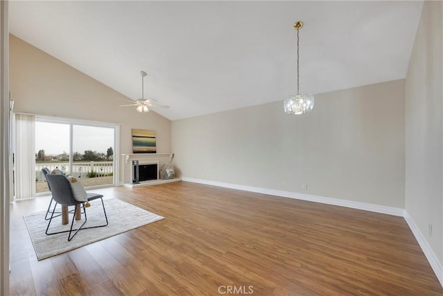 unfurnished living room featuring a fireplace with raised hearth, wood finished floors, baseboards, high vaulted ceiling, and ceiling fan with notable chandelier