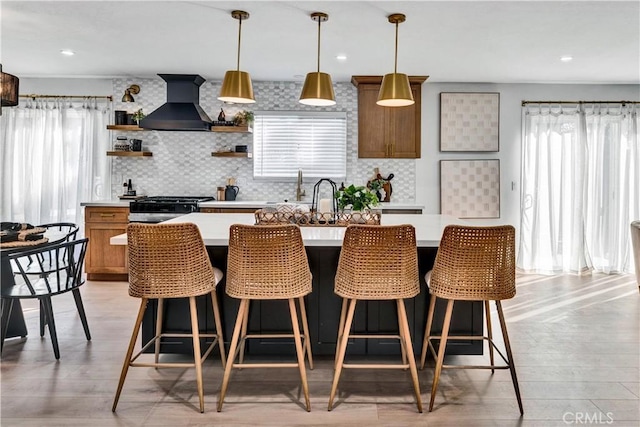 kitchen with wall chimney range hood, open shelves, brown cabinets, decorative backsplash, and light countertops