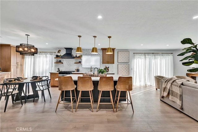 kitchen with an island with sink, light countertops, backsplash, open shelves, and wall chimney exhaust hood