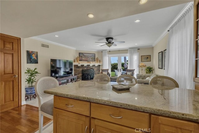 kitchen with crown molding, open floor plan, light stone counters, ceiling fan, and a stone fireplace