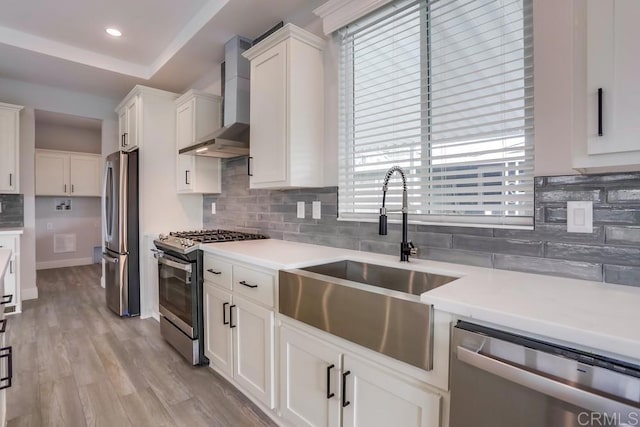 kitchen with white cabinets, stainless steel appliances, a sink, backsplash, and wall chimney range hood