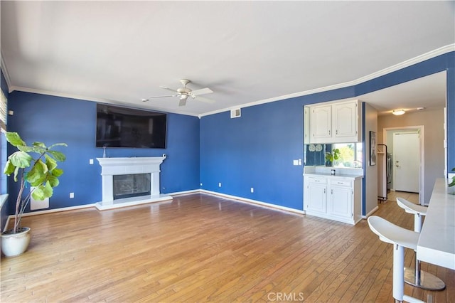 unfurnished living room featuring ceiling fan, light wood finished floors, crown molding, a glass covered fireplace, and baseboards