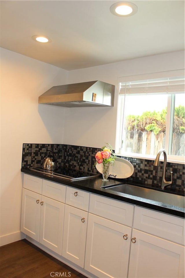 kitchen featuring wall chimney range hood, decorative backsplash, black electric stovetop, white cabinets, and a sink