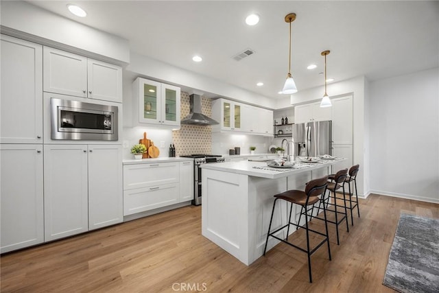 kitchen featuring wall chimney range hood, white cabinetry, light wood-style flooring, appliances with stainless steel finishes, and a kitchen bar