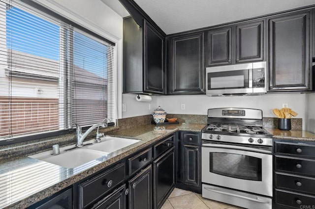 kitchen with stainless steel appliances, dark cabinetry, light tile patterned floors, a sink, and dark stone counters