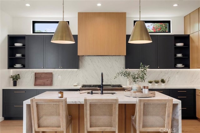kitchen with light stone counters, open shelves, a sink, pendant lighting, and a kitchen island with sink
