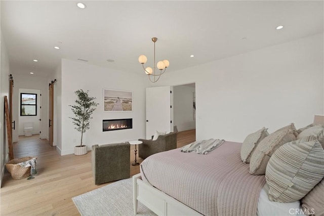 bedroom featuring light wood finished floors, a barn door, a glass covered fireplace, recessed lighting, and a notable chandelier