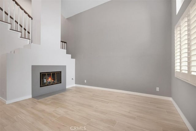 unfurnished living room featuring stairway, a high ceiling, wood finished floors, baseboards, and a glass covered fireplace