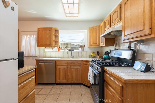 kitchen with tile counters, under cabinet range hood, a sink, tasteful backsplash, and appliances with stainless steel finishes