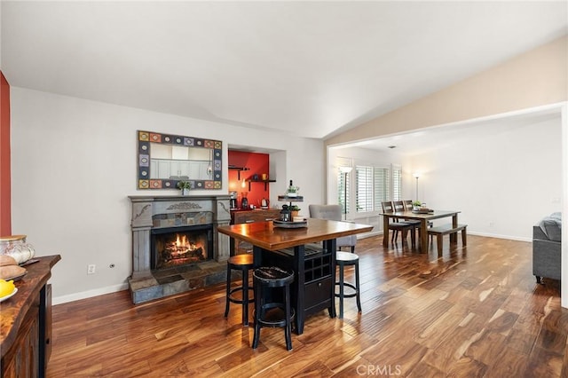 dining room featuring baseboards, wood finished floors, a warm lit fireplace, and vaulted ceiling