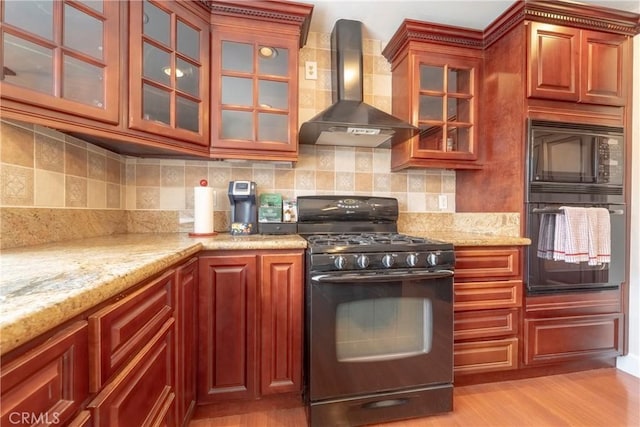 kitchen featuring wall chimney range hood, light stone counters, light wood-style floors, glass insert cabinets, and black appliances
